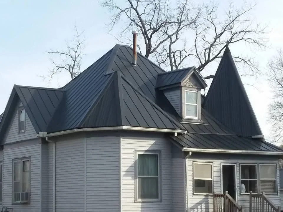 Charcoal metal standing seam roof with dormers, brick chimney, and white vinyl siding